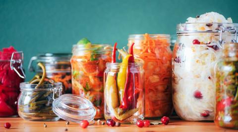 Image of various fermenting foods in jars over a blue background and sitting on a wooden table, including peppers, carrots, etc.