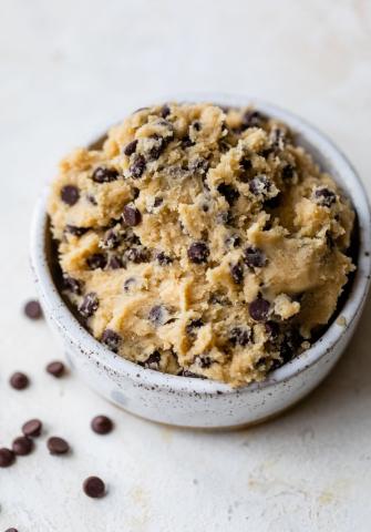 Edible cookie dough in a ceramic dish with a few loose chocolate chips on the white marble countertop beneath it.