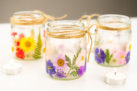 Image of the craft featuring small clear jars with twine wrapped around the rim of the jar with colorful dried flowers pressed and glued on the inside of the jar. 
