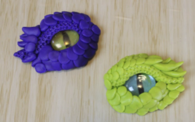 One purple and one green dragon eye made out of clay resting on a wood table.