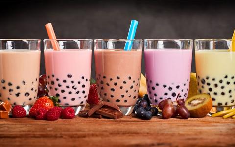 5 glasses of bubble tea next to one another, the first with caramel in front of it, the second with strawberries, the third with chocolate, the fourth with blueberries, and the last with kiwis.