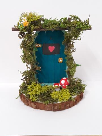 Image of the craft featuring a painted wooden door and wooded platform with moss and embellishments to make it look like a fairy door in the woods. 