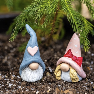 Two painted gnomes, one with a blue hat and a pink heart on it, and another with blonde braids, a pink hat and a red bow, both sat outside on dirt with pine needles hanging above them.