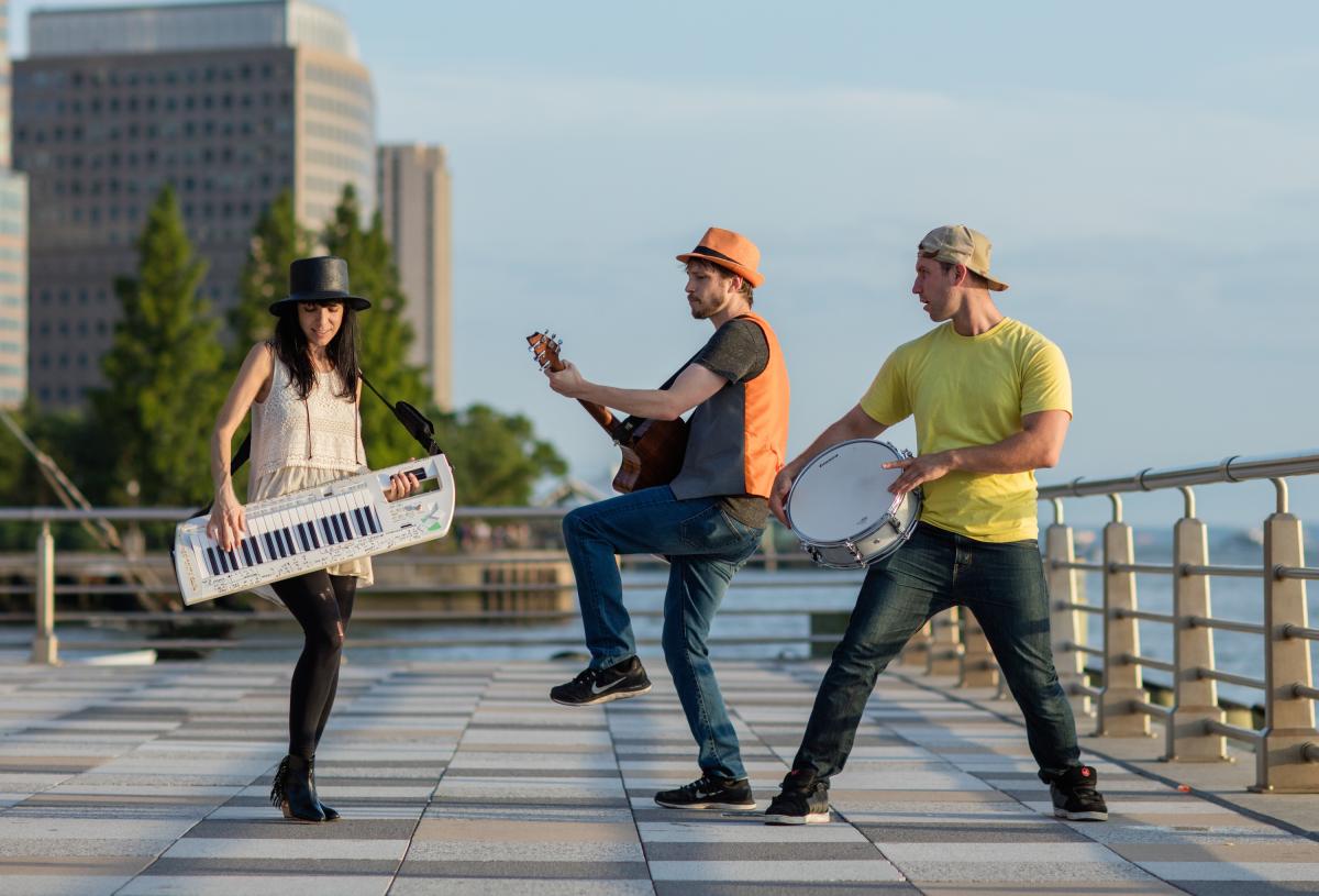 Image of 3 people on a rooftop in the city. The woman is playing a keytar (Keyboard guitar) one man is playing the guitar and another a snare drum. 