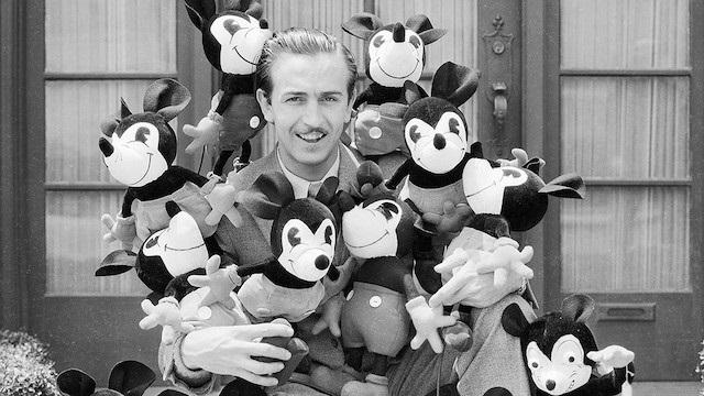 Black and white image of Walt Disney as a young man holding 8 stuffed Mickey Mouse plushes, smiles on their faces. Two seem to be on the ground beside him as well, as he stands in front of closed doors.