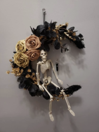 Image of a spooky moon hanging wreath on a wall made of black, orange, and yellow flowers shaped like a moon that a skeleton sits on