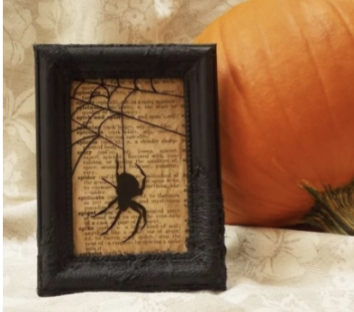 Image of a picture frame with newspaper in the middle and a black spider and web silhouette over it. The frame is on off white table cloth and in front of a pumpkin. 